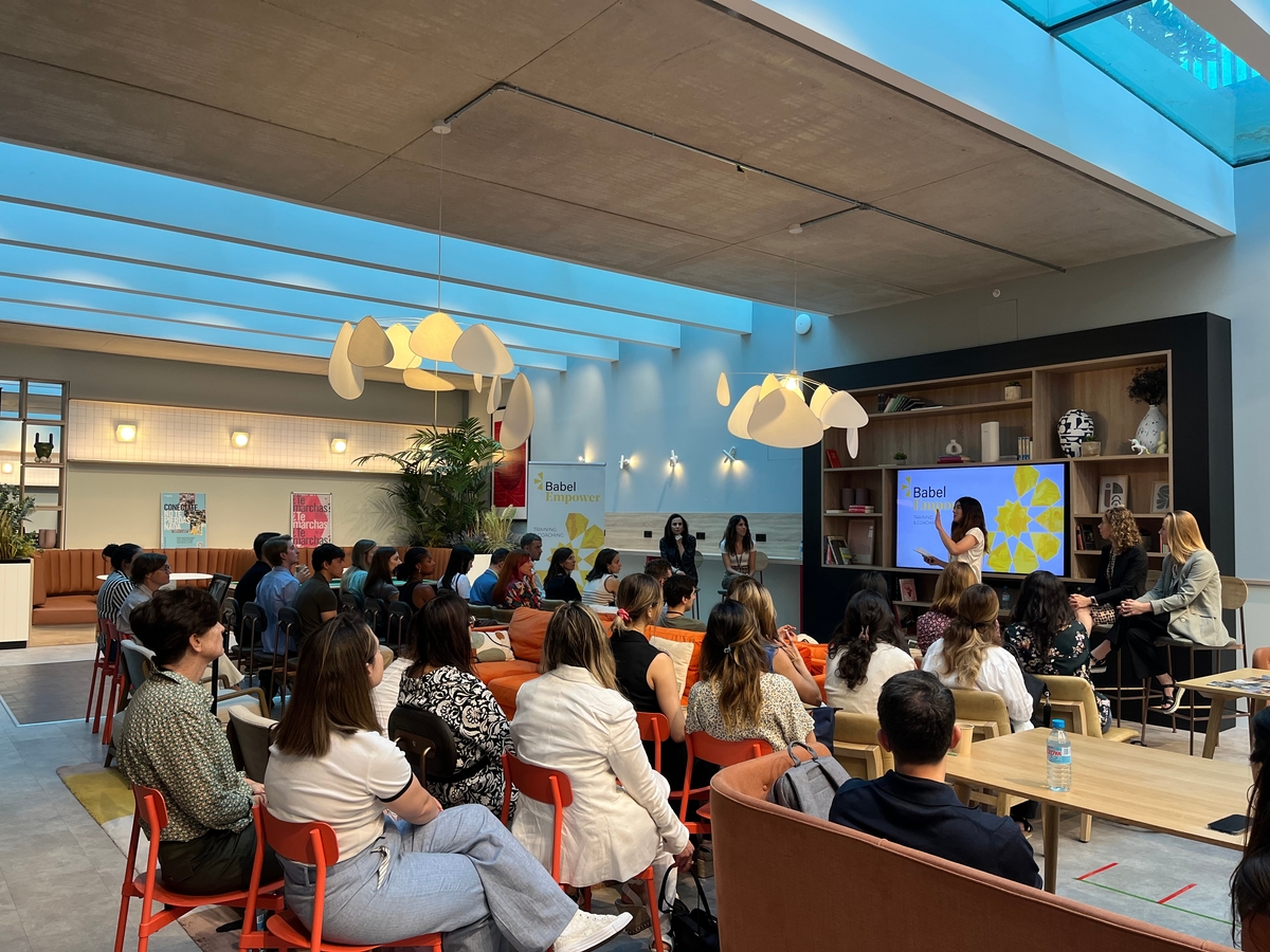 A group of people seated in a spacious, modern room with a high ceiling, attending a presentation. A panel of speakers is in front of a screen displaying the word "Babel" and a design featuring yellow flowers. The room is filled with natural light.