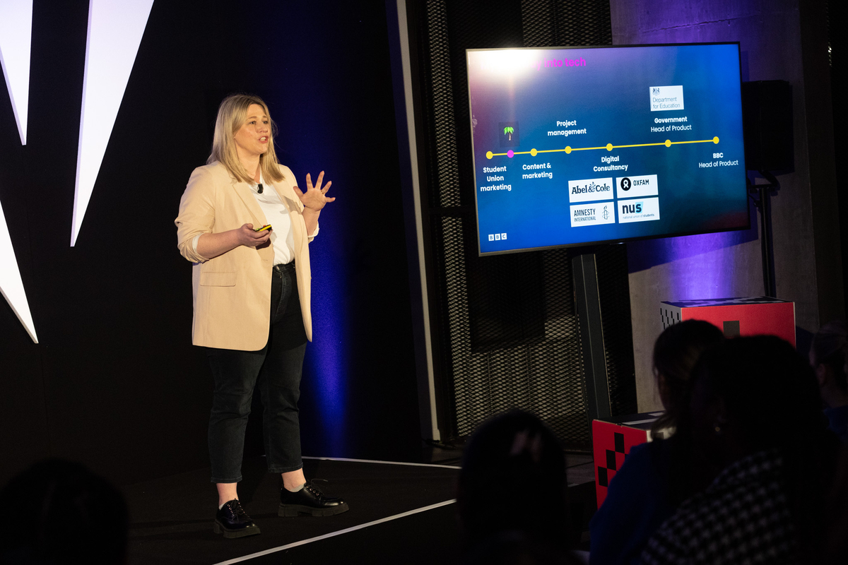 A person on stage giving a presentation, gesturing with one hand. They stand beside a screen showing a career timeline with logos like BBC and NUS. The audience is seated, watching attentively.