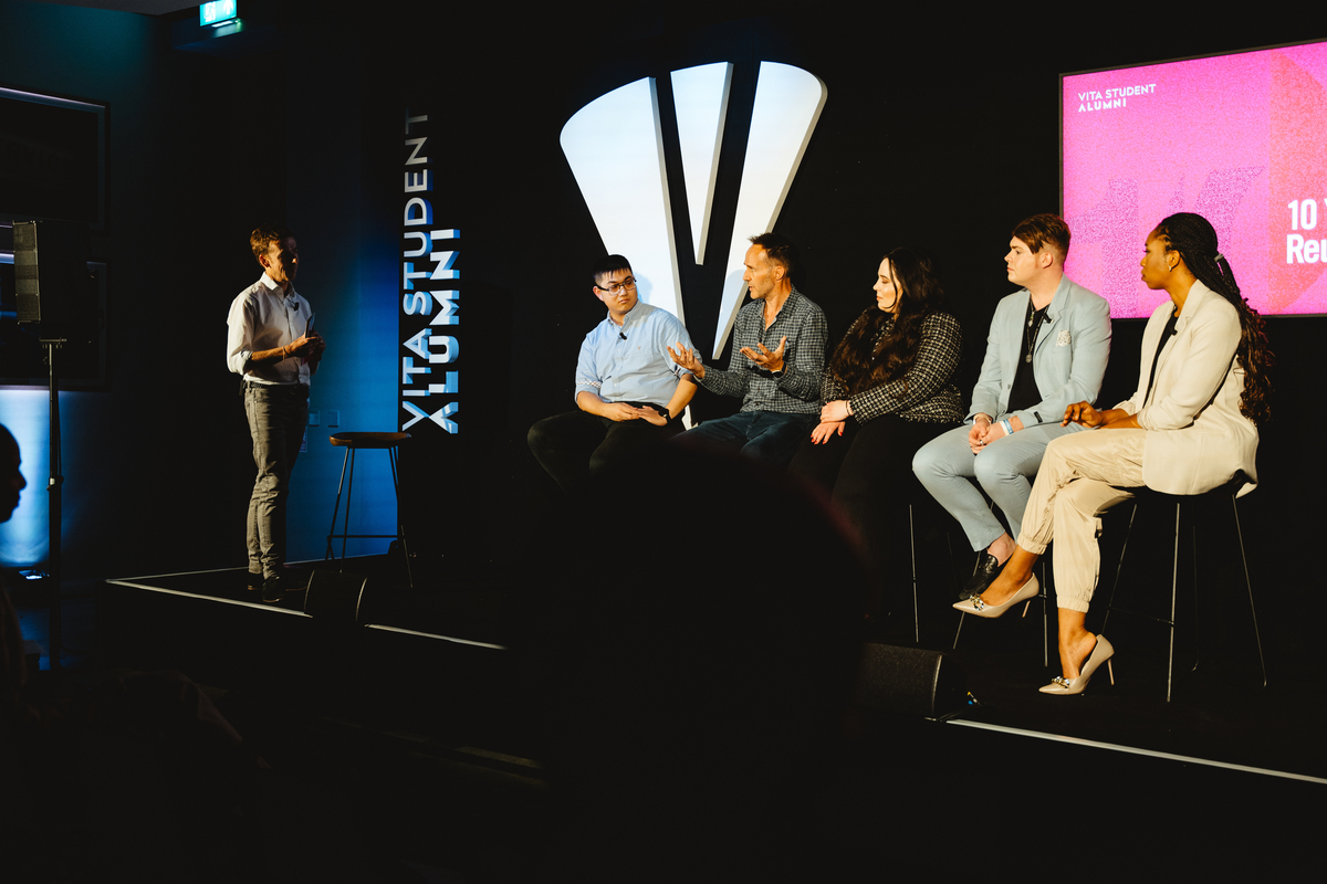 A person stands at a podium addressing a panel of five individuals seated on stage. The background features a screen with text and the logo "Vita Student Alumni." The setting is dimly lit with a focus on the panelists.
