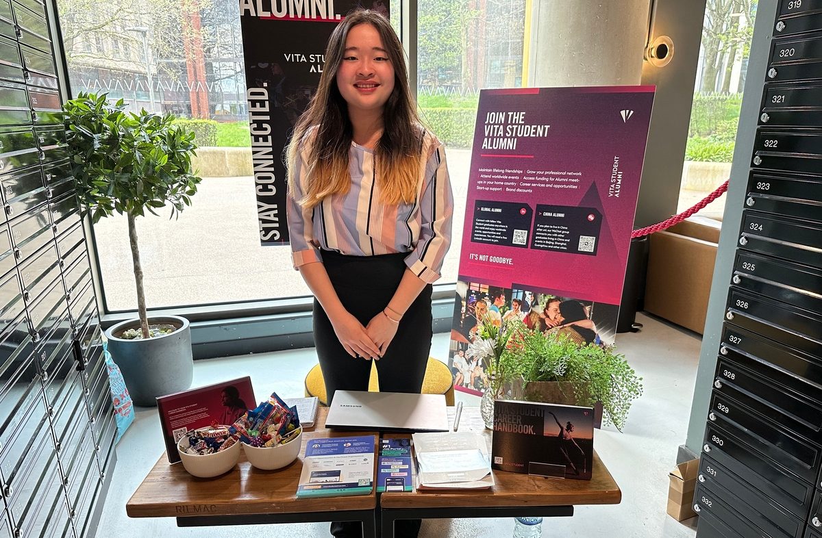 A woman stands behind a table with informational flyers, a laptop, a bowl of candy, and a poster about joining the Vita Student Alumni. The setting includes plants and a window showing an outdoor view.