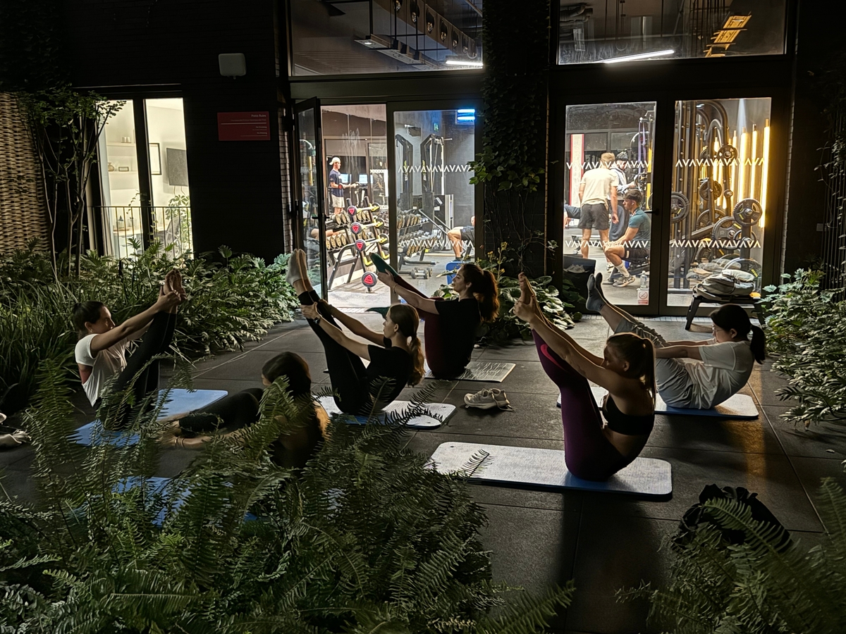 A group of people practice yoga on mats in an outdoor garden setting at night. They are performing a pose with their legs raised, holding their feet, surrounded by lush green plants. Indoors, others can be seen working out in a gym with equipment.
