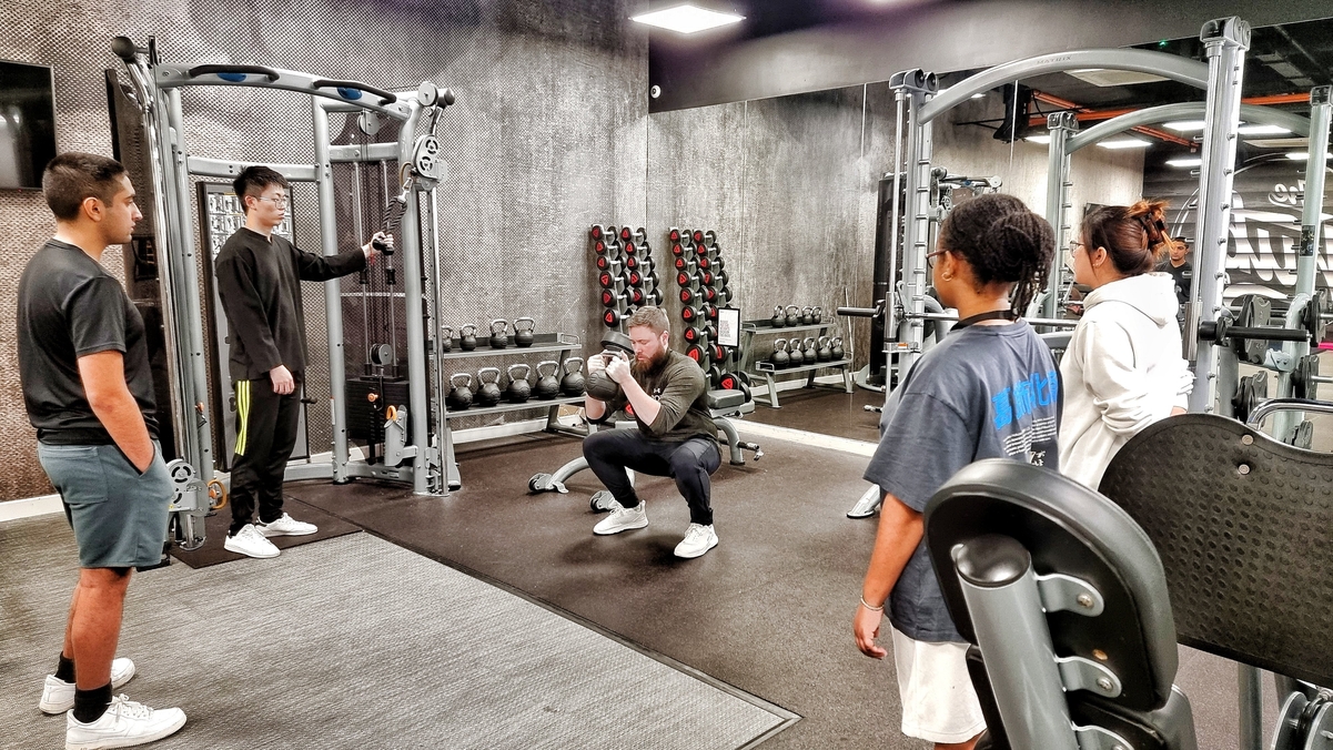 Five people are in a gym. One person is doing squats while the others observe. Various gym equipment, like dumbbells and machines, are in the background. The setting is modern and well-lit.