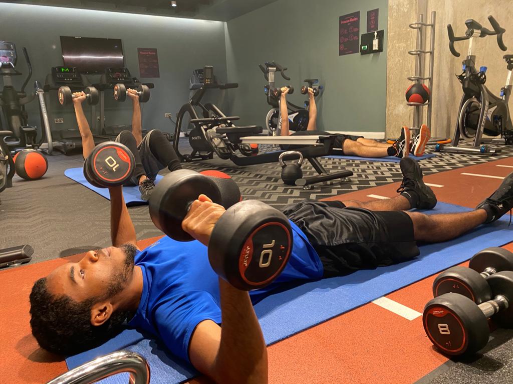 A person in a blue shirt lies on a gym mat lifting dumbbells. Exercise equipment, including spin bikes and a treadmill, are visible in the background. Another person is stretching on a mat. The setting suggests a gym environment.