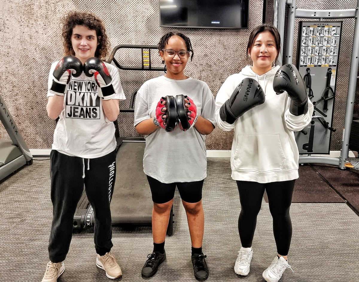 Three people stand in a gym wearing boxing gloves. The person on the left wears a "New York DKNY Jeans" shirt, the person in the center holds focus mitts, and the person on the right wears a hoodie. They are in front of exercise equipment.