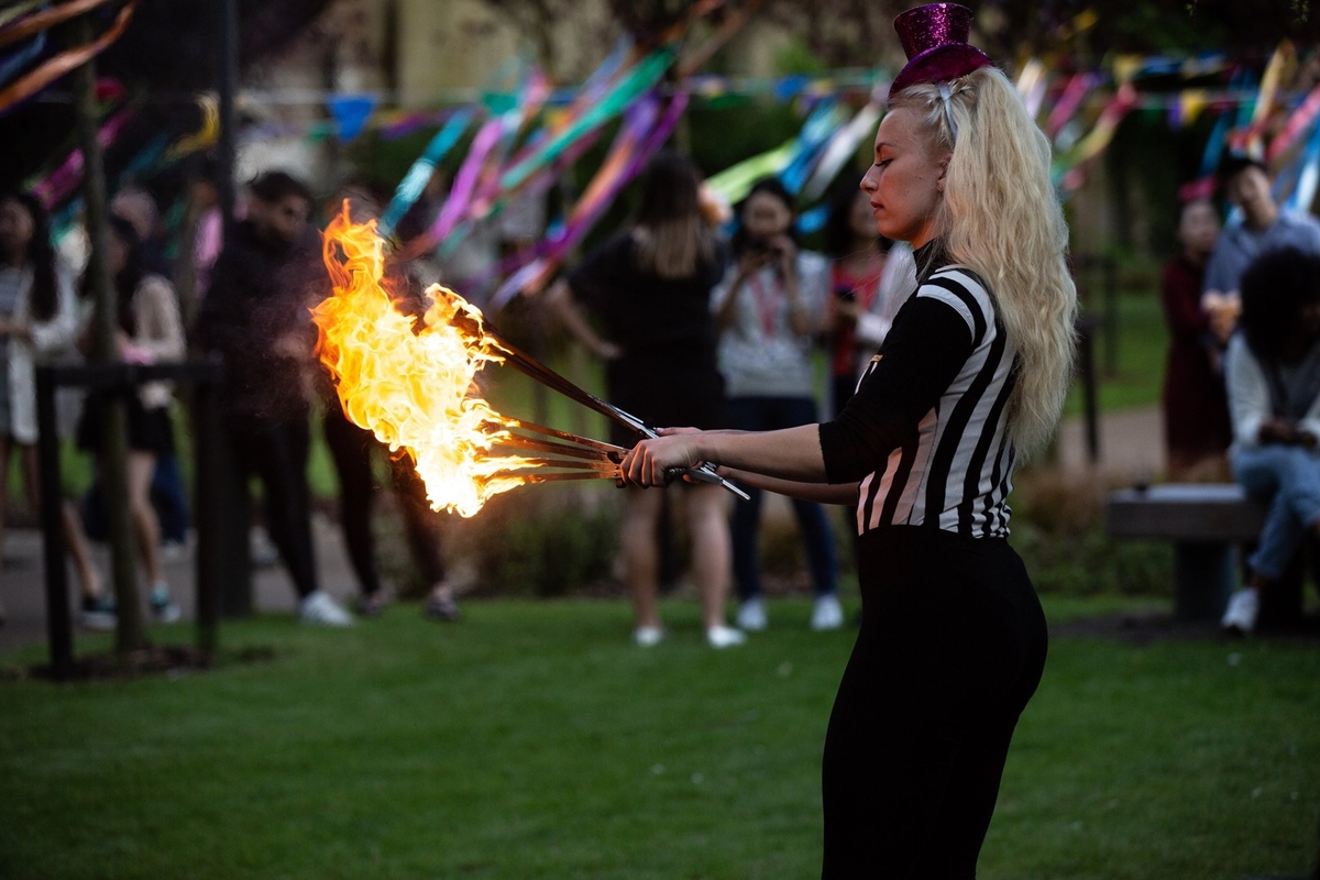 A performer with long blonde hair wearing a striped top and a small hat balances two flaming batons in a park. Colorful ribbons and a crowd of people are in the background.