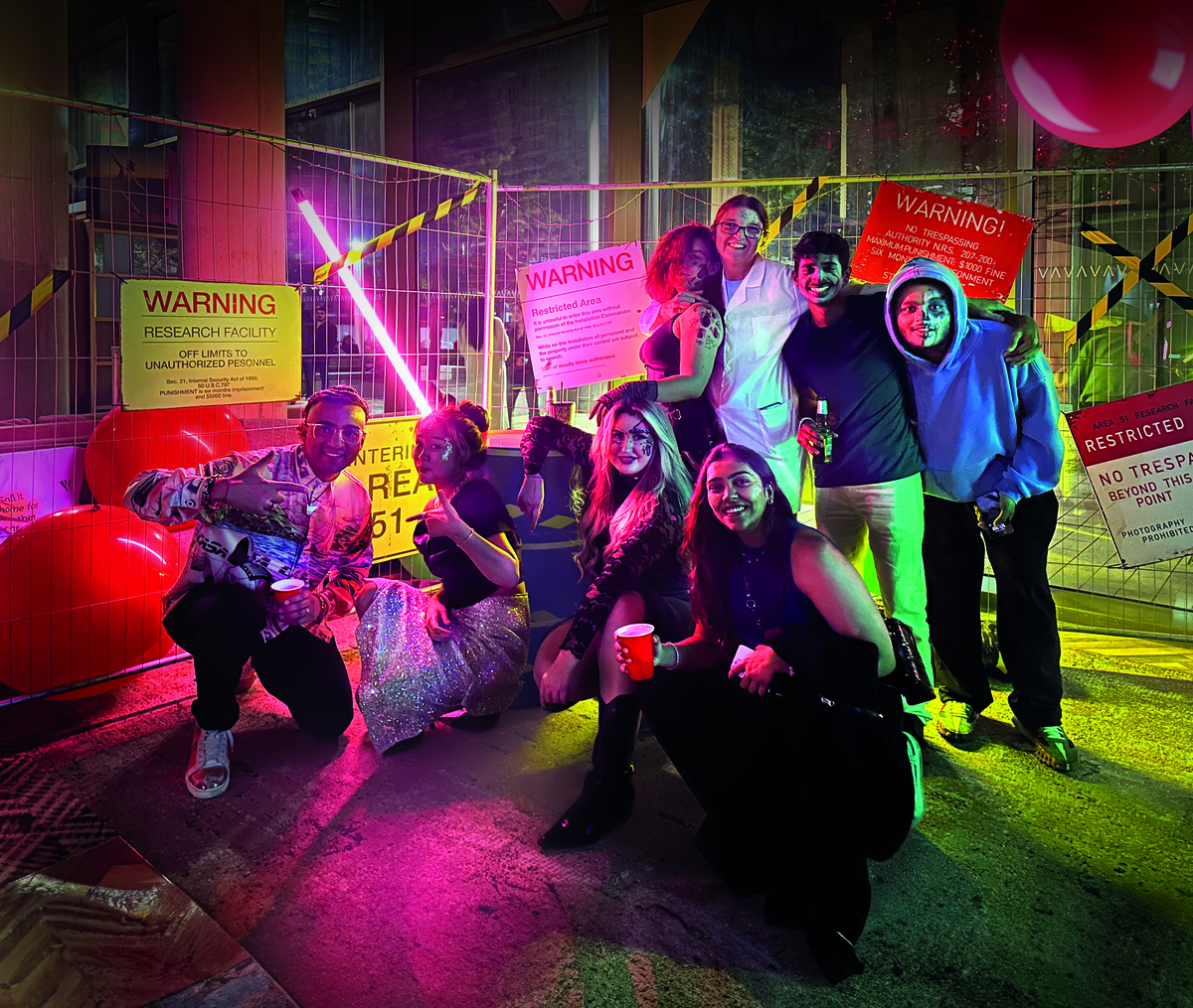 A group of people pose and smile at a colorful, themed party with "Warning" signs and caution tape in the background. They are holding drinks and balloons, surrounded by vibrant lights.