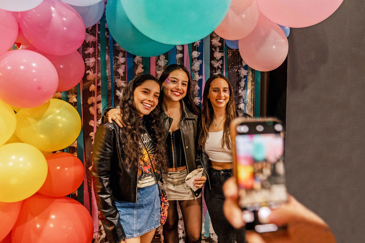 Three women pose for a photo at a colorful party. They stand in front of a backdrop with balloons in pink, yellow, and blue. One woman in a leather jacket holds a phone capturing the moment.