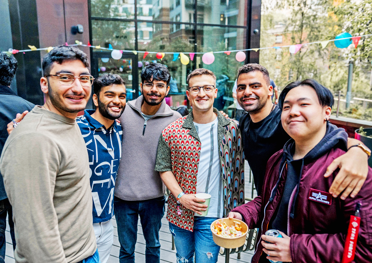 A group of six people smiling outdoors at a gathering. One holds a bowl of snacks, and another has a drink. Colorful bunting decorates the background, and there are buildings and trees visible behind them. They're standing on a wooden deck.