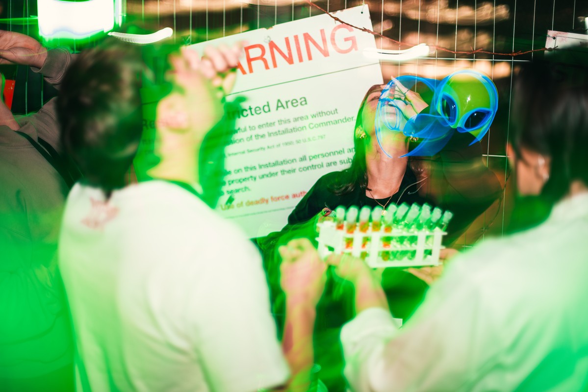 A group of people in a party setting with green lighting. One person wears a blue alien mask, holding test tubes in a rack. A warning sign is partially visible in the background. The scene appears lively and surreal.
