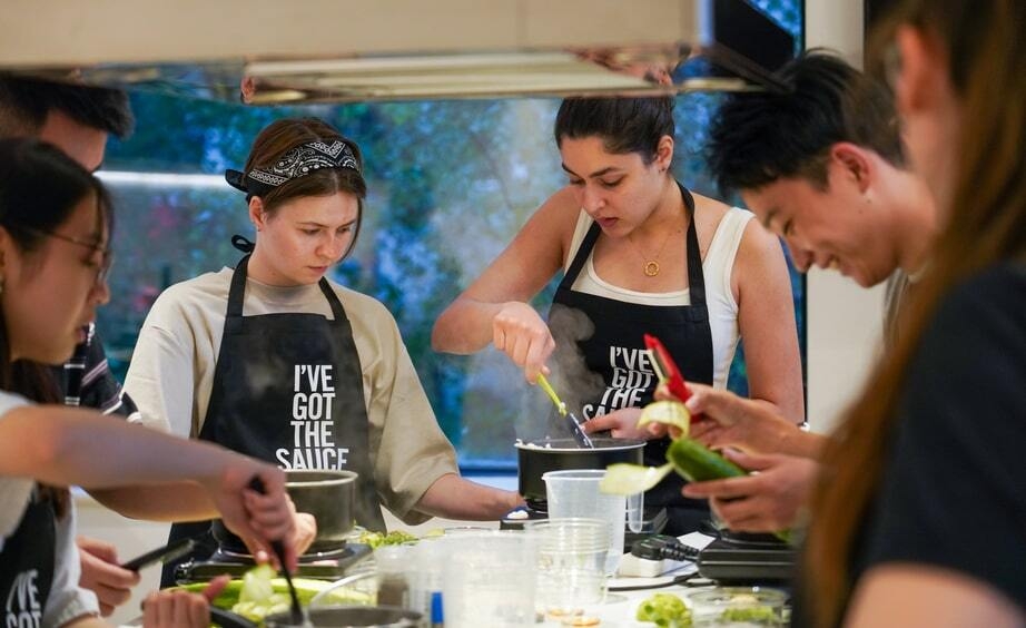 A group of young adults wearing aprons is cooking together in a kitchen. One person stirs ingredients in a pan while others focus on their tasks, like chopping vegetables. The atmosphere is lively and collaborative.