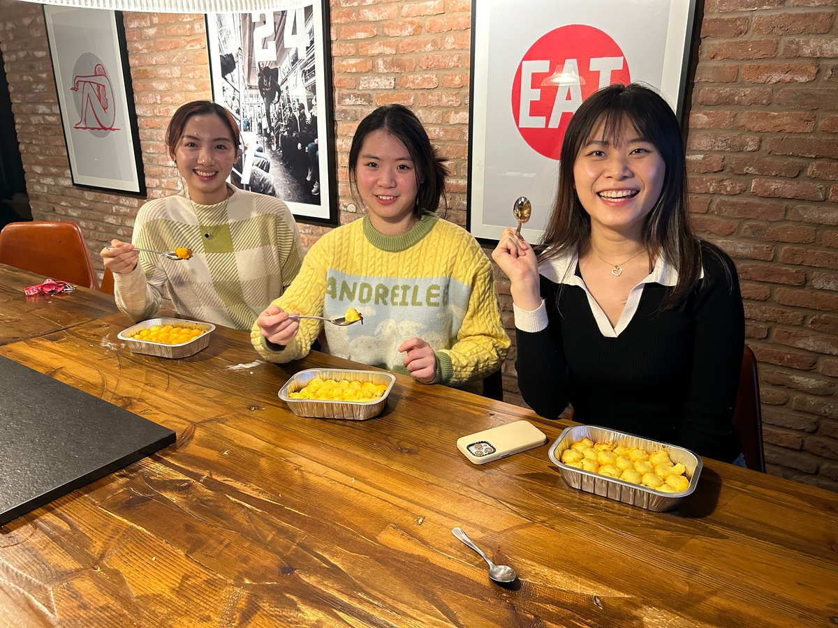Three people sitting at a wooden table, smiling, and holding spoons above trays of food. Behind them are brick walls and framed artwork. The setting appears cozy and casual.