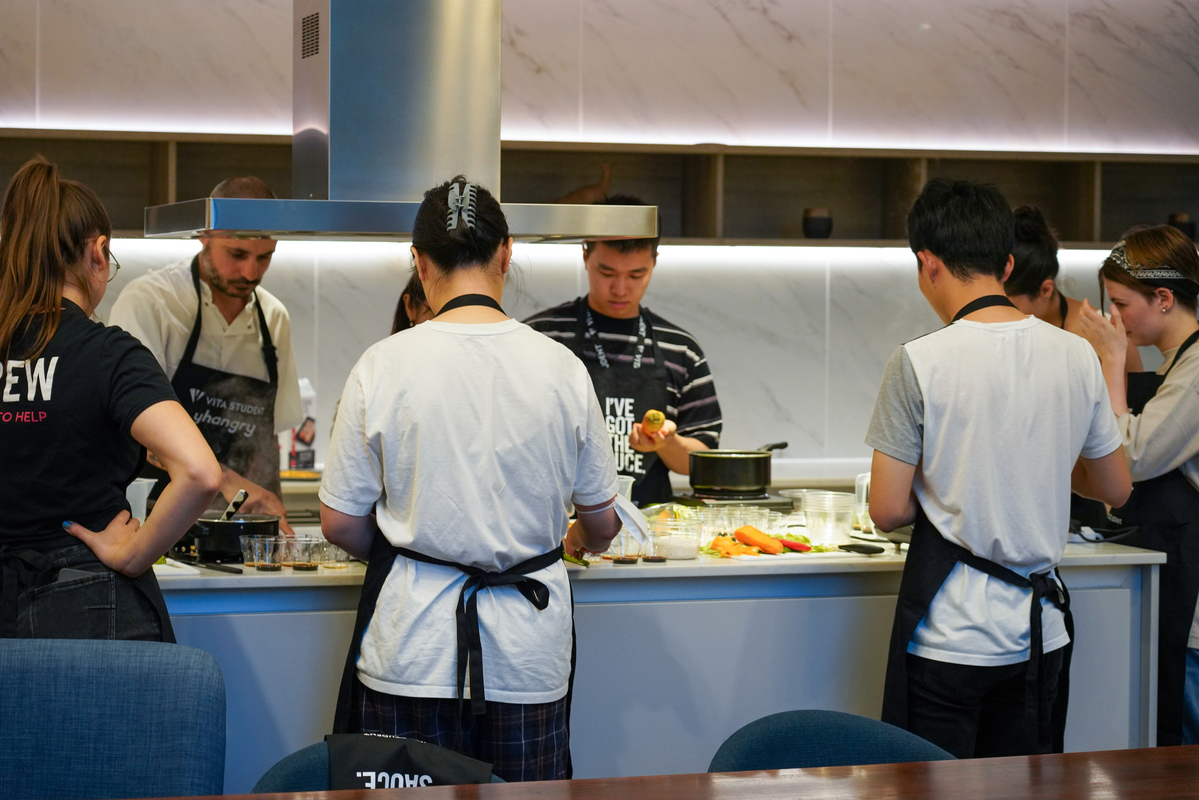 A group of people cooking together in a modern kitchen. They are wearing aprons and preparing various ingredients on the counter. Some are chopping while others are cooking at the stove. The atmosphere is focused and collaborative.