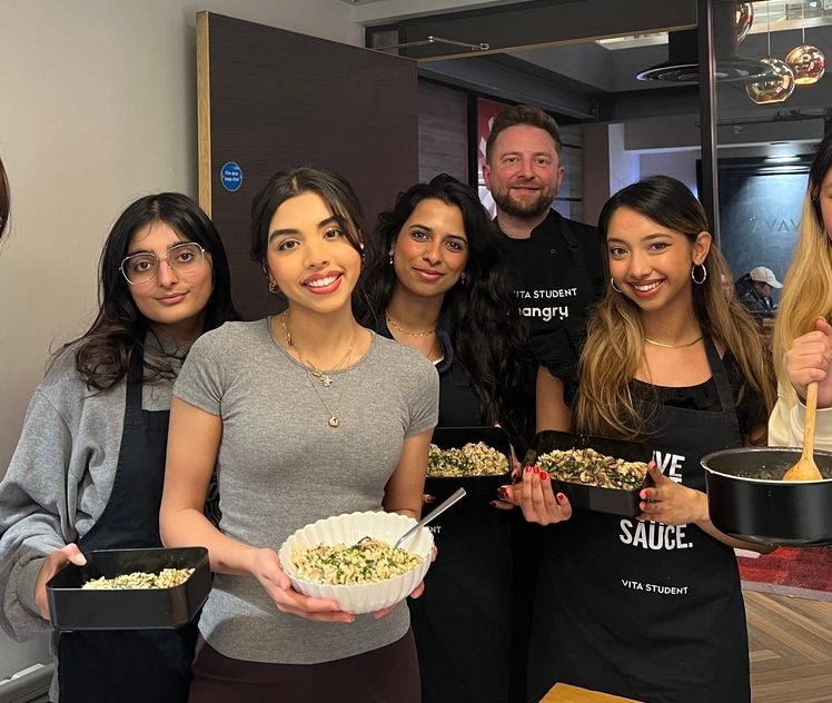 A group of six people holding bowls of salad, standing in a kitchen. Some are wearing black aprons with logos. They are smiling and appear to be participating in a cooking activity.
