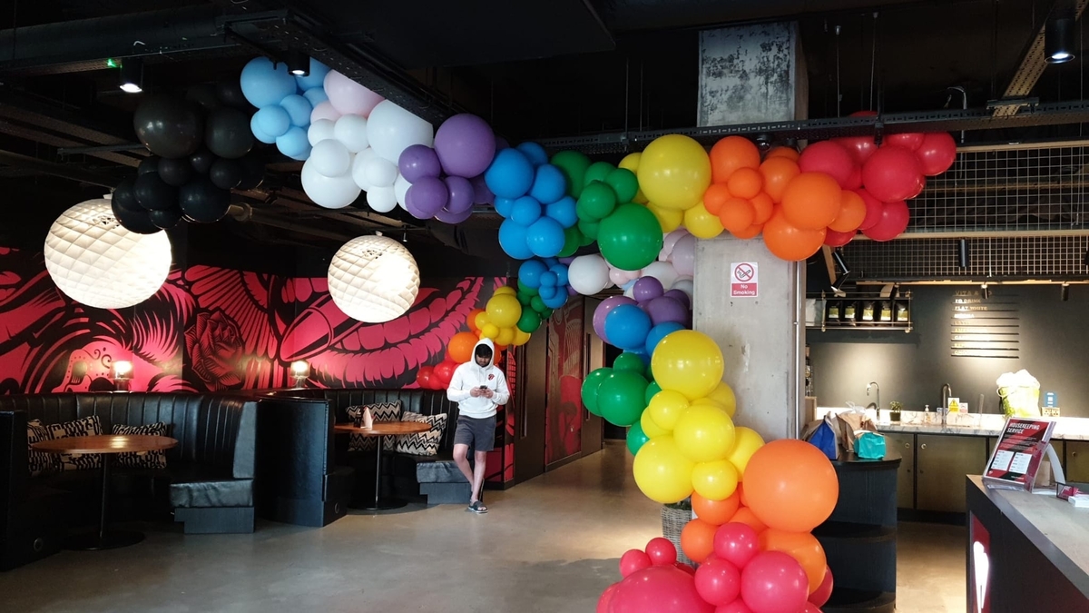 A colorful archway of balloons in various colors, including black, white, purple, blue, green, yellow, orange, and red, adorns a modern indoor space. A person stands beneath the arch, and seating areas with round lights are visible in the background.