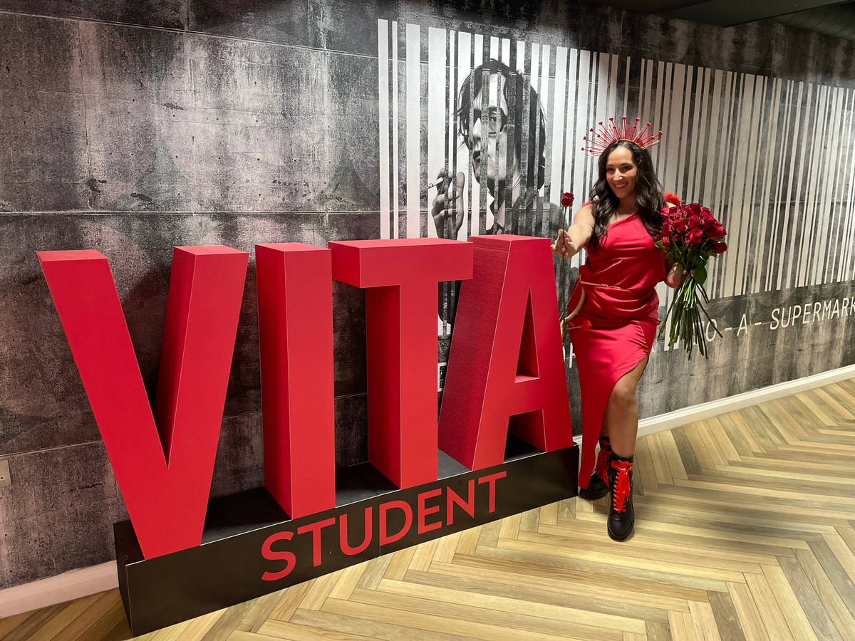 A person in a red dress and boots, holding roses, stands next to large red letters spelling "VITA Student". There is a black-and-white mural in the background.