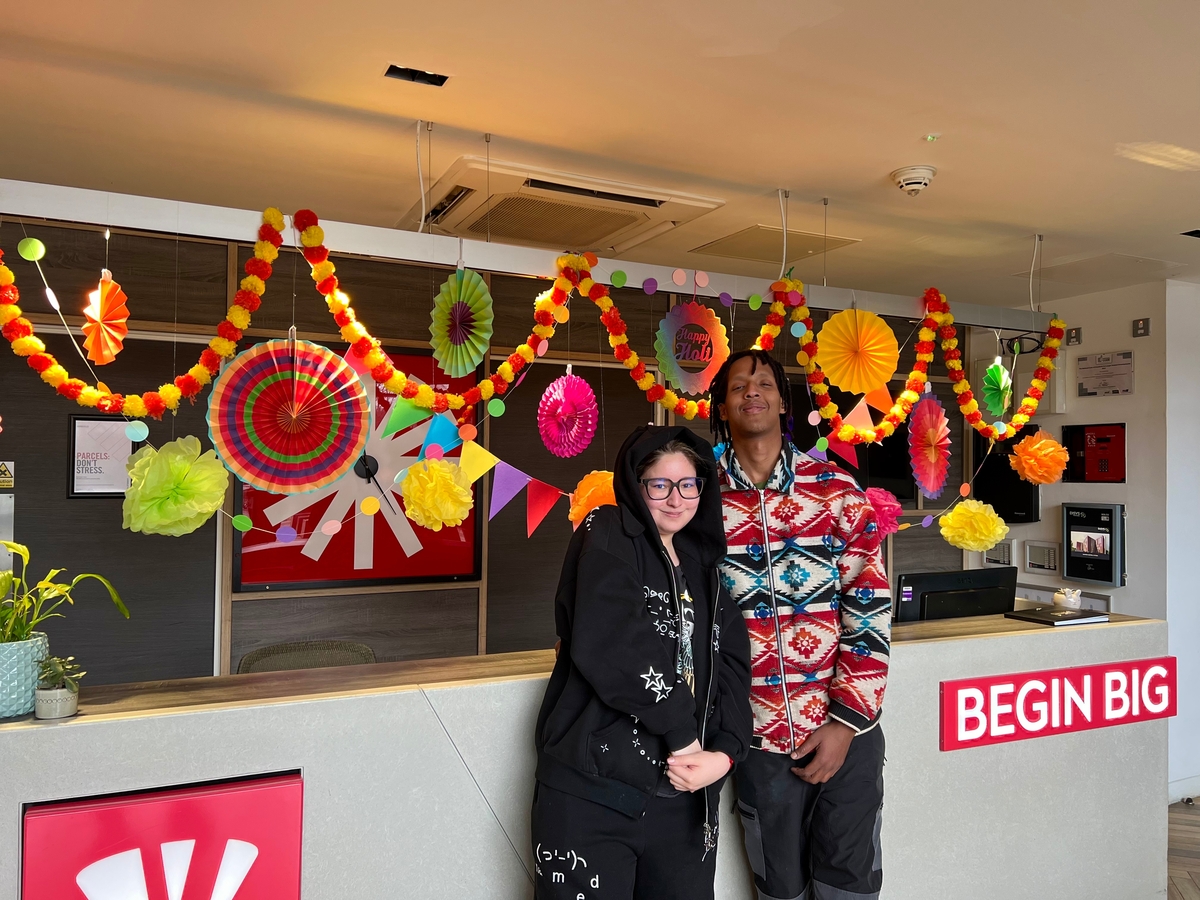 Two people standing in front of a decorated reception desk with colorful paper fans and garlands. The desk has a sign that reads "BEGIN BIG." A few potted plants and a microwave are visible in the background.