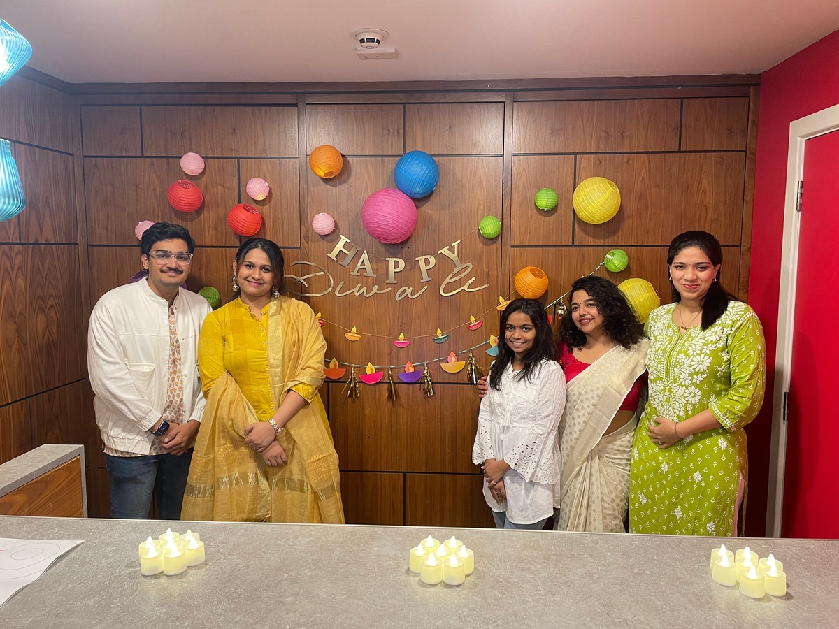 Five people stand together smiling in front of a "Happy Diwali" sign with colorful lanterns and string lights. They are dressed in festive Indian attire. The background features wood paneling, and LED candles are placed on the counter.