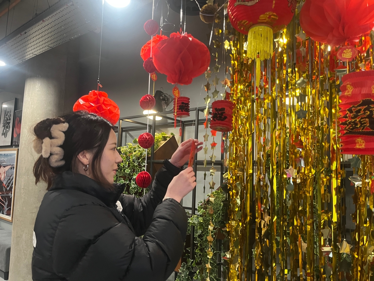 A person decorates a space with red lanterns and golden tinsel for a festive occasion. The surroundings feature greenery and other decorations, creating a lively atmosphere.