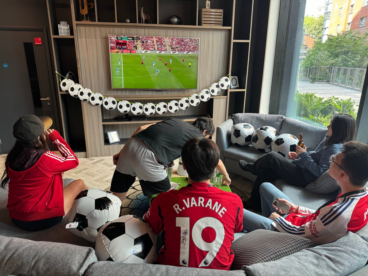 A group of people in a living room watching a soccer match on a wall-mounted TV. They are sitting on couches decorated with soccer ball pillows. One person is in front of the TV, preparing food. Soccer-related decorations are visible around the room.