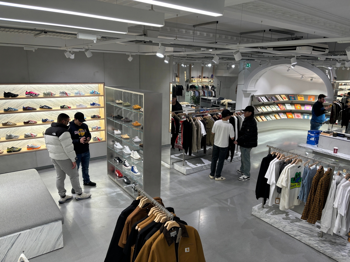 A trendy clothing store with a modern design. Customers browse racks of clothing, and shelves display various sneakers. The interior features sleek grey flooring and bright, even lighting. Pictures are displayed on the wall in the background.