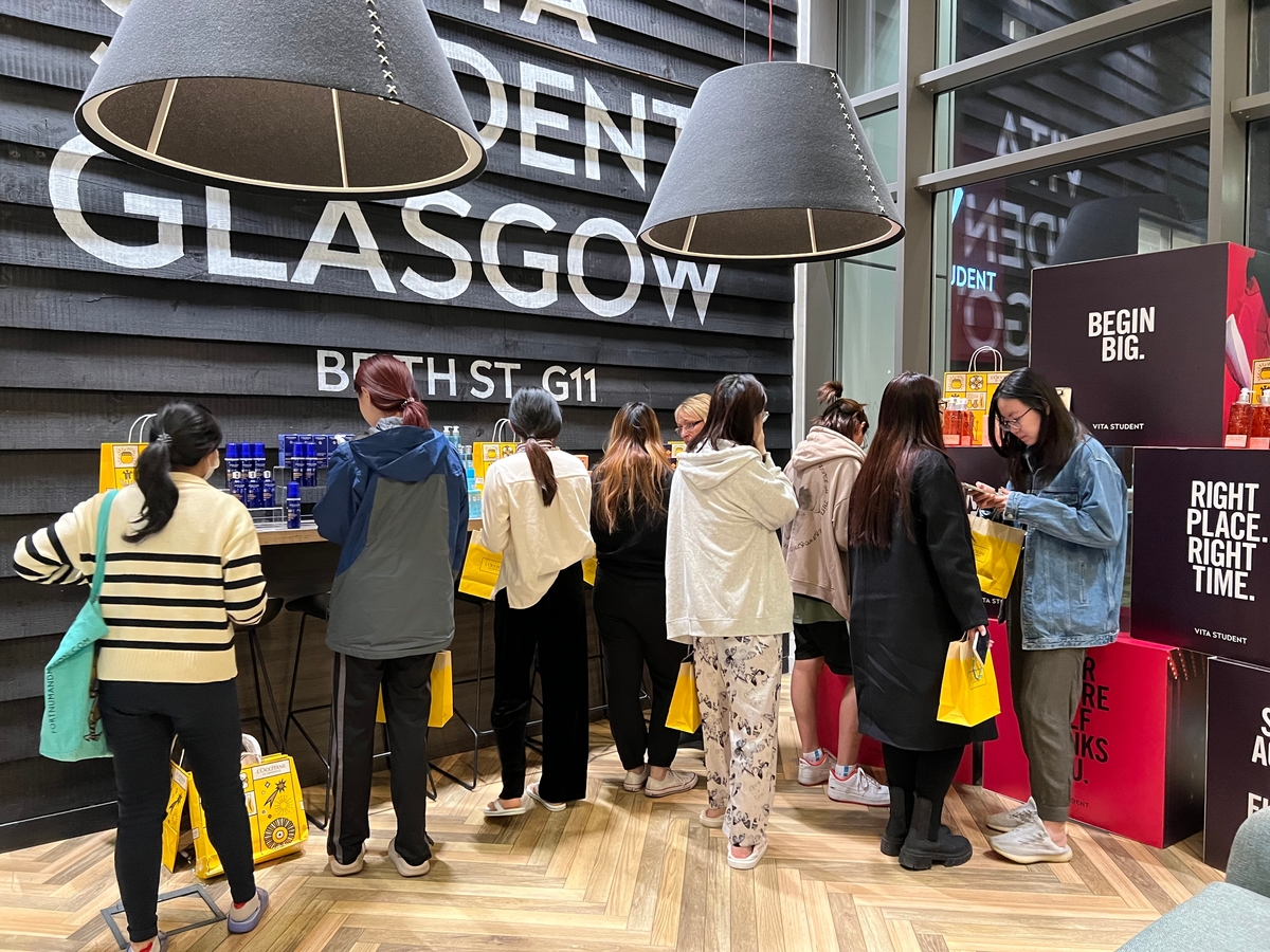 A group of people stands at a counter inside a store. They are browsing products while holding yellow bags. The store has large, modern pendant lights and a wall displaying the text "Glasgow." Red and white boxes are stacked nearby.