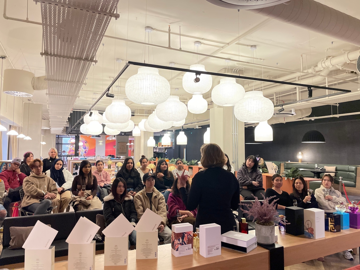 A person is speaking to a seated audience in a modern room with hanging lights. People are seated at tables with various items placed on them, including white boxes and potted plants. The decor features industrial-style ducts and cozy furniture.