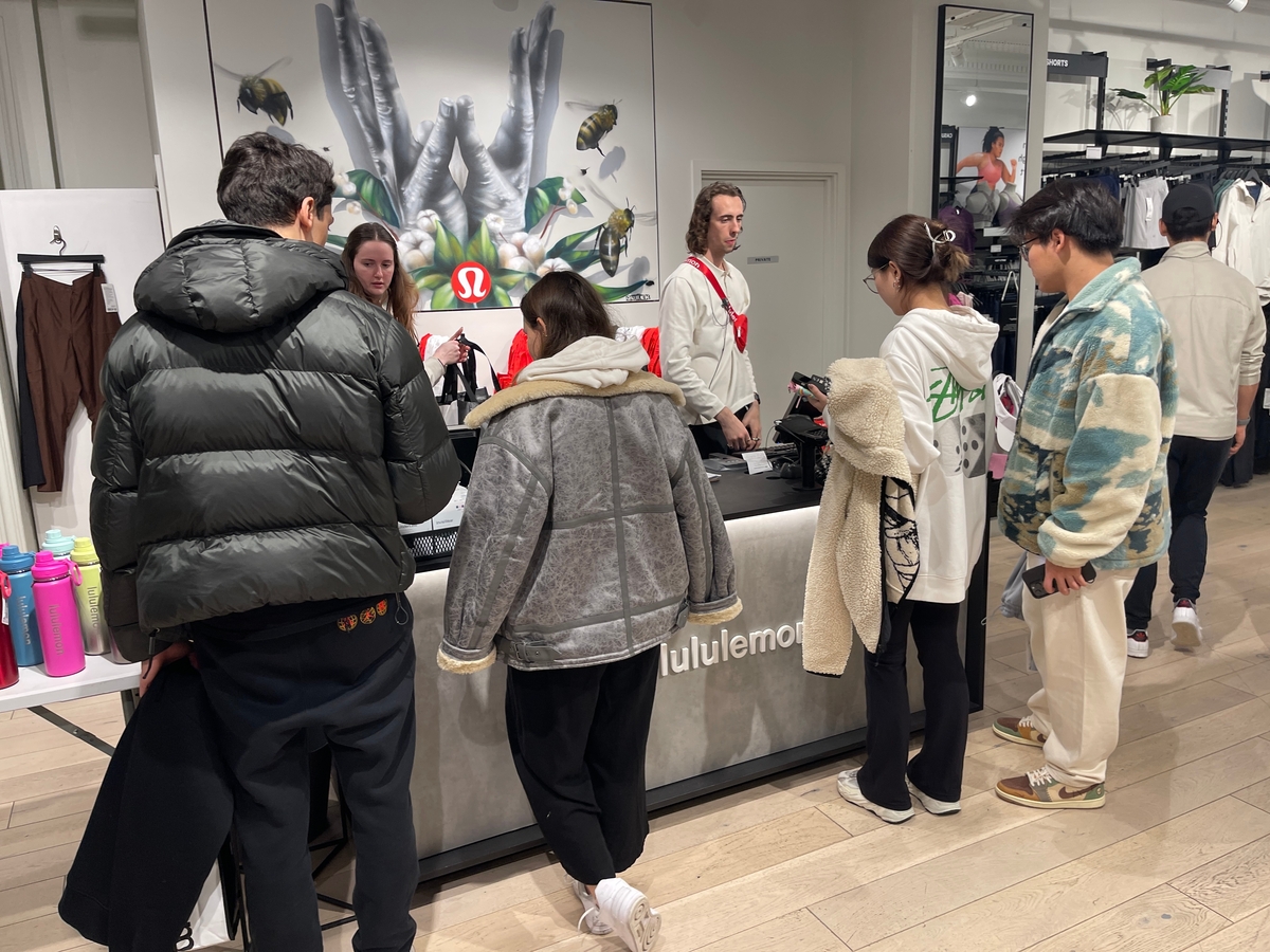 Customers standing at a Lululemon counter inside a store. A group of people are engaged in conversation with employees. Several items, including brightly colored water bottles, are displayed nearby. Clothing and accessories are visible in the background.