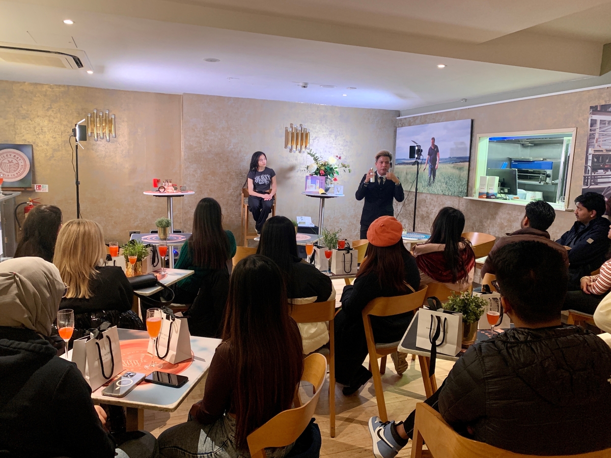 A group of people seated in a room listen to two speakers in front. The setting has a casual ambiance, with a few tables, potted plants, and beverages. The walls have framed pictures, and various items are displayed on a table near the speakers.