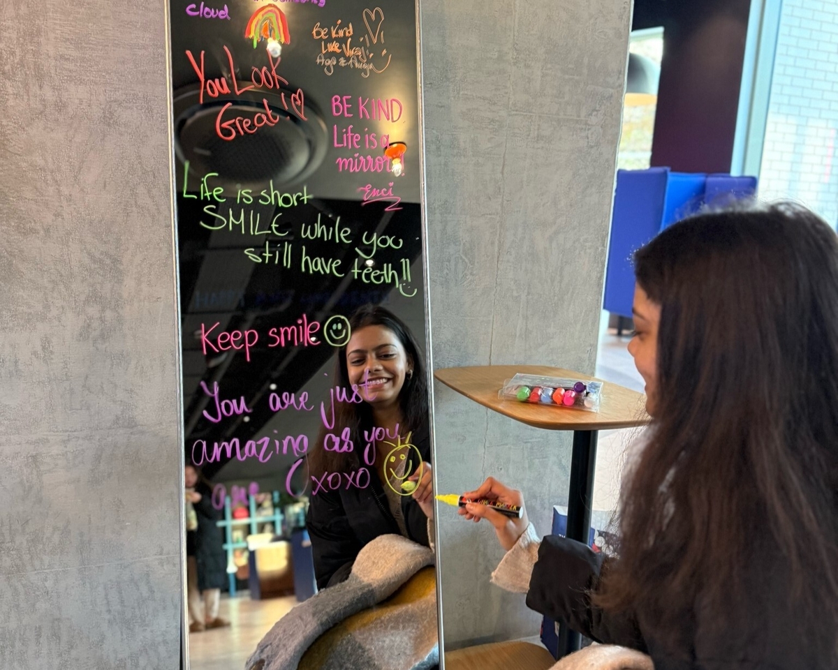 A woman smiling at her reflection while writing with colorful markers on a mirror, adorned with motivational messages and drawings. A small table with more markers is beside her.