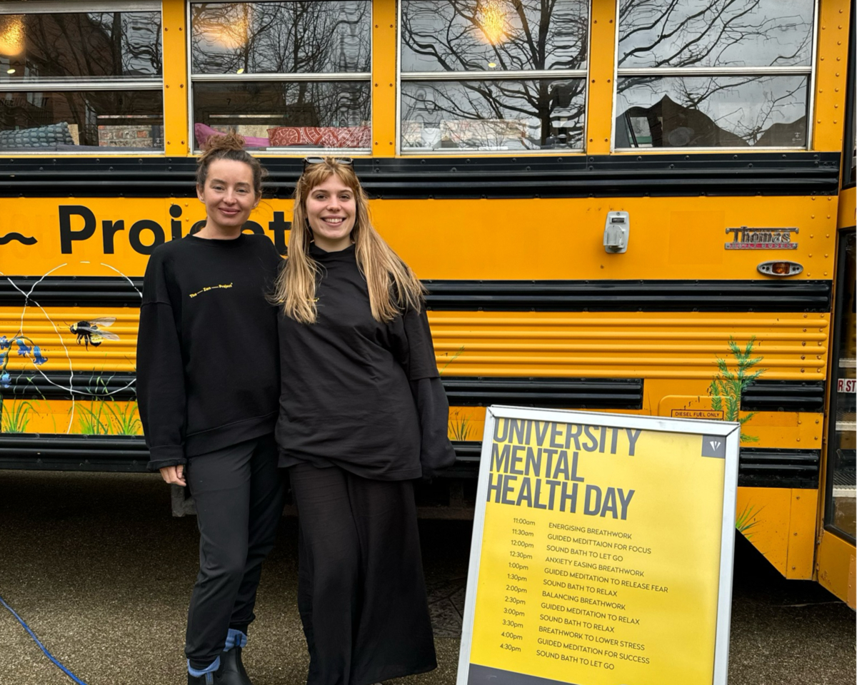 Two people wearing black outfits stand smiling in front of a yellow bus with "University Mental Health Day" displayed on a schedule board. The bus has a mural, and trees are reflected in its windows.