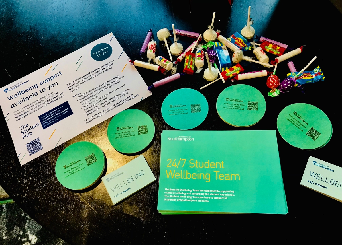 A table displays wellbeing support flyers, wellness coasters, a brochure titled "24/7 Student Wellbeing Team," and an assortment of colorful lollipops and candies. The materials emphasize student support services.