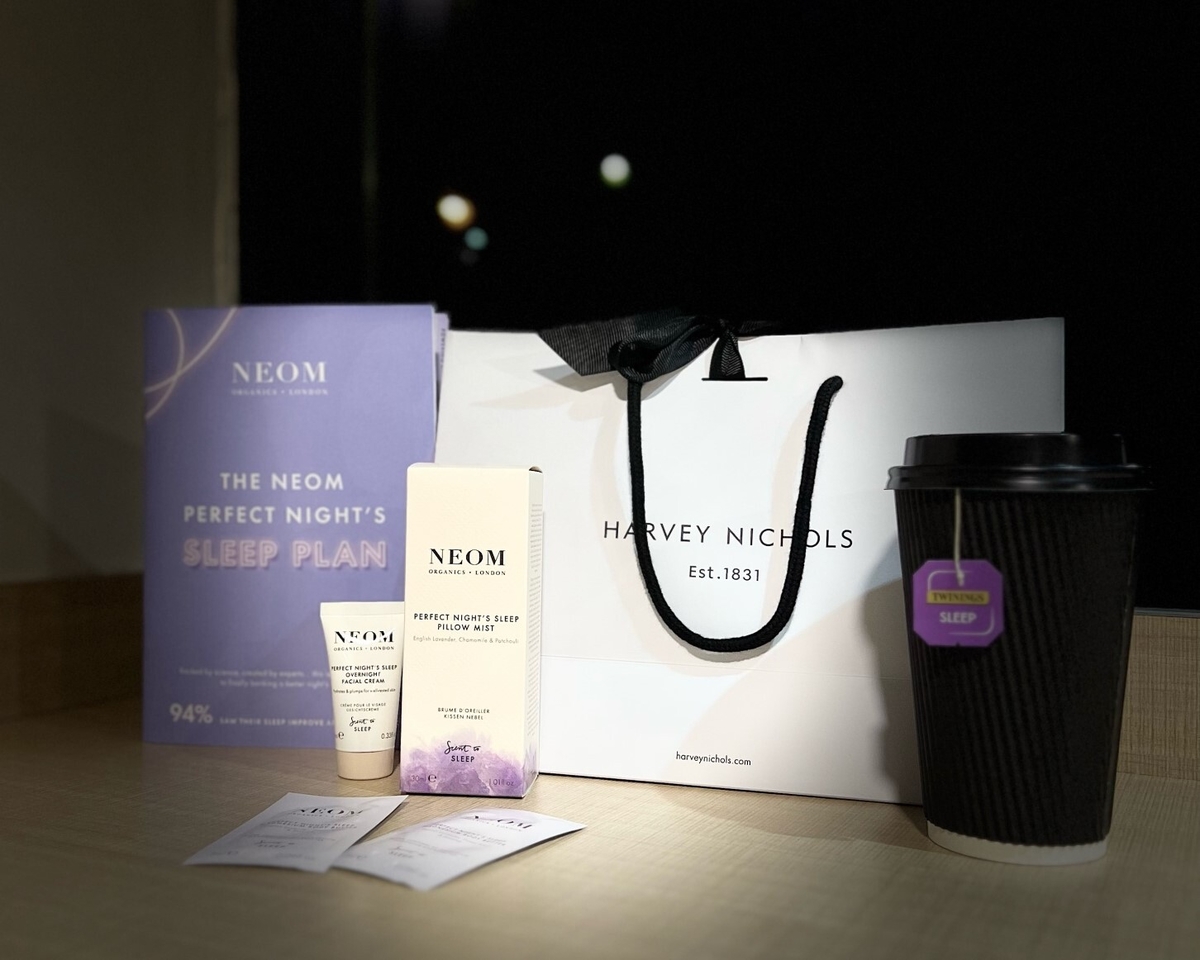 A table displays a Harvey Nichols shopping bag, a purple NEOM sleep plan booklet, a black coffee cup with a "sleep" tag, and two NEOM product boxes along with sample sachets. The background shows a blurry nighttime window view.