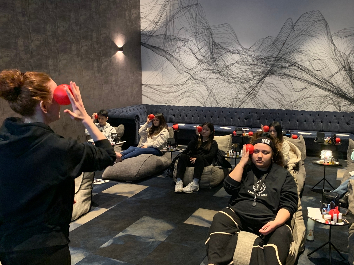 A group of people relax in a dimly lit room with modern decor. They are seated on cushions and wearing black eye masks. A person at the front holds a red object up to their face, leading the session.