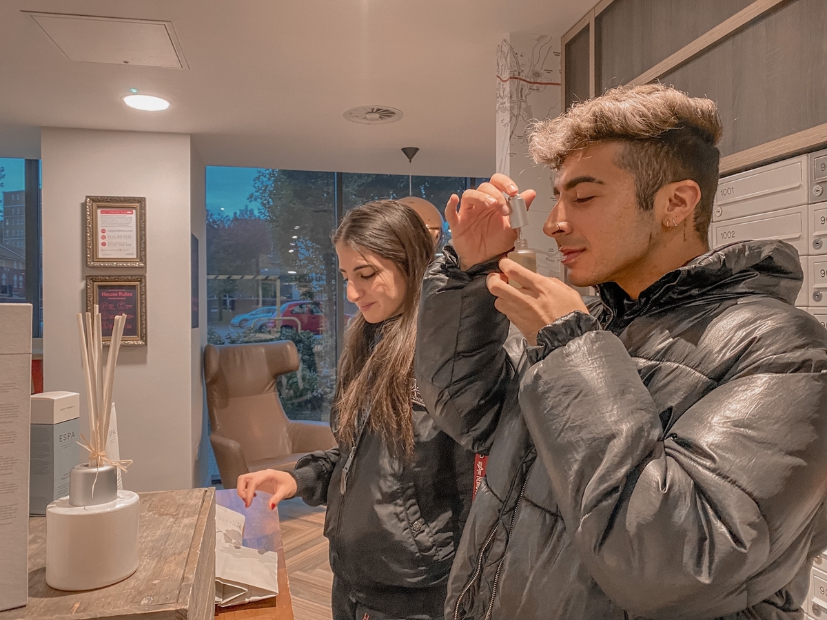 Two people stand indoors near a table. One person sniffs a scent stick from a container of reed diffusers, while the other person observes with a smile. They are both wearing dark jackets, and a blurred street view is visible through the window.