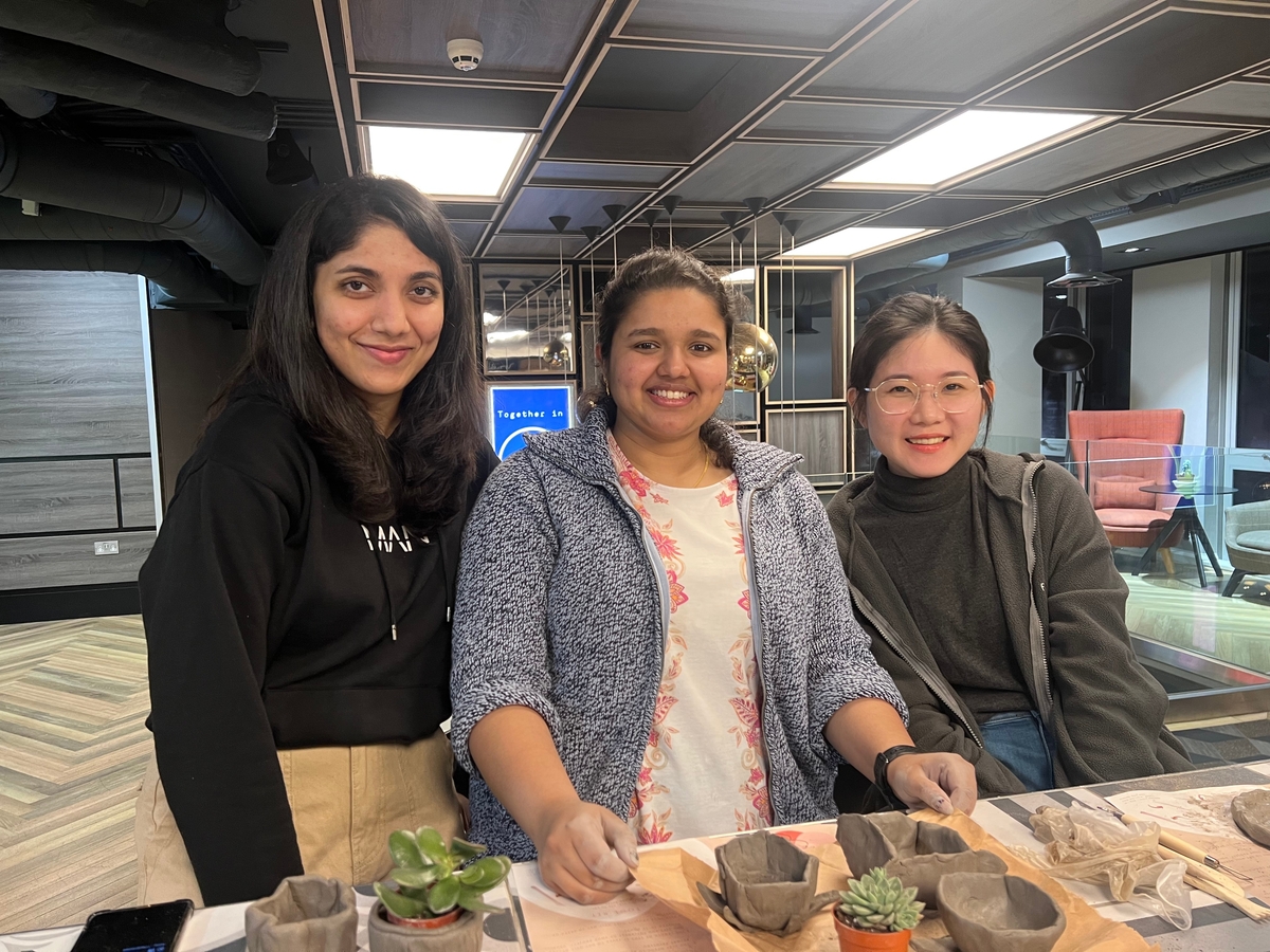 Three people smile while seated at a table with small plants and crafting materials. The setting appears to be indoors, with a modern design featuring wood accents and overhead lighting.