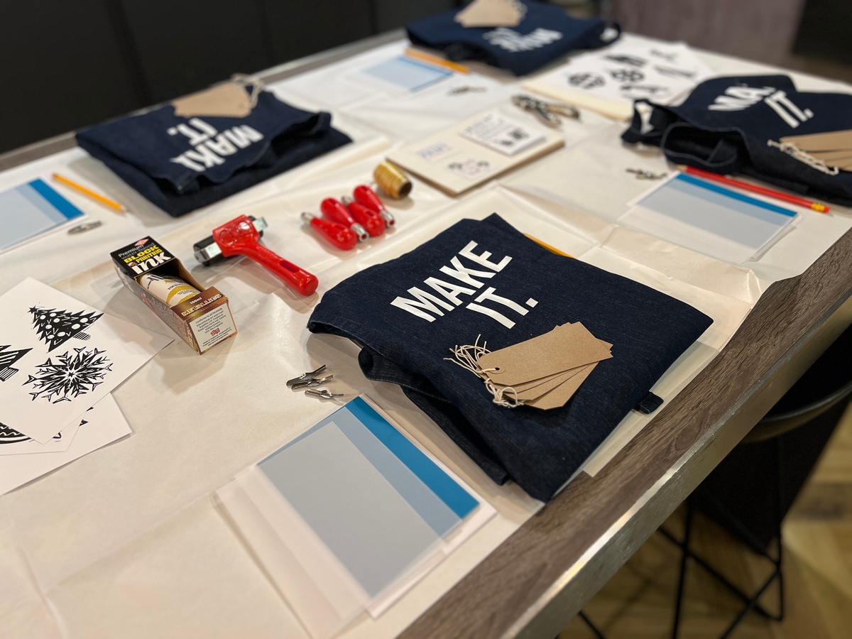 A crafting workshop table set with denim aprons, stencil designs, tags, fabric markers, scissors, a red hot glue gun, and a box of Tikki markers. The aprons have "MAKE IT." printed on them.