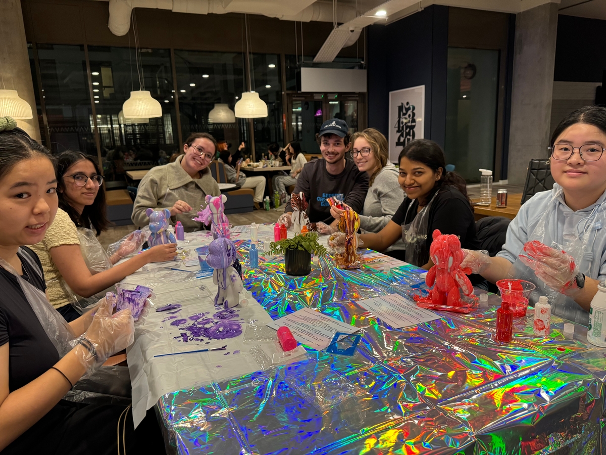 A group of people sitting around a table covered in a shiny, iridescent tablecloth, painting ceramic figures. Various colors and art supplies are visible. They are wearing gloves and some have aprons. The setting appears to be a casual, indoor space.