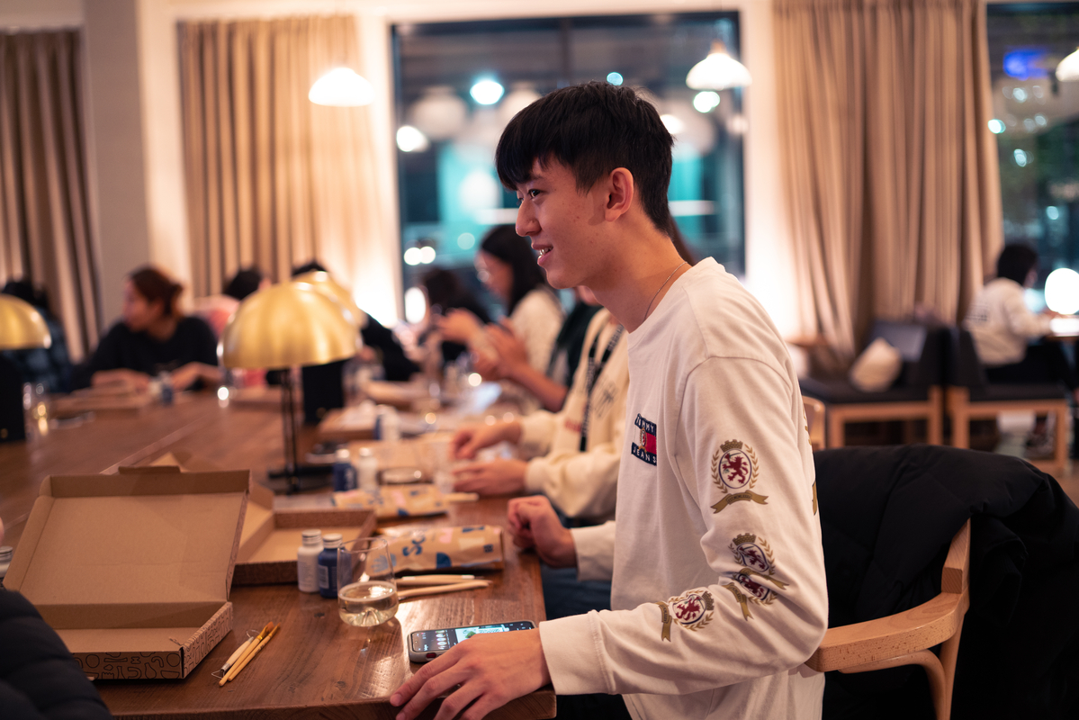 A young man in a white long-sleeve shirt sits at a wooden table in a cozy restaurant, smiling. There are pizza boxes and drinks on the table. Other diners in the background are engaged in conversations. Warm lighting and curtains add to the ambiance.