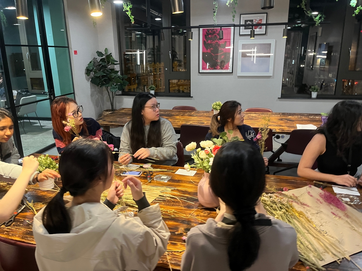A group of people seated around a large wooden table, engaged in a flower arranging activity. The room is well-lit with large windows and modern decor, including hanging plants and framed art on the walls.