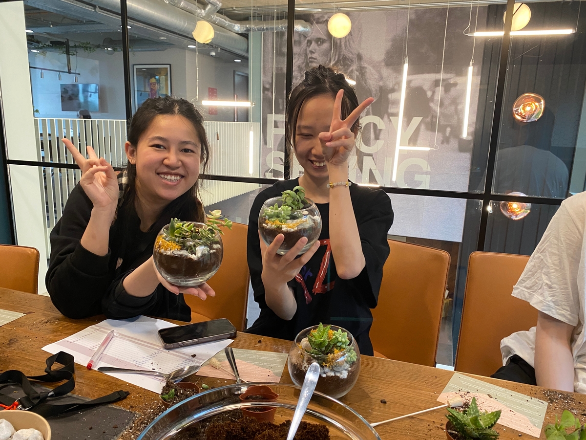 Two people smiling and holding small terrariums in a room. One is making a peace sign with their hand. Papers and crafting materials are on the table. A blurred poster is visible in the background.