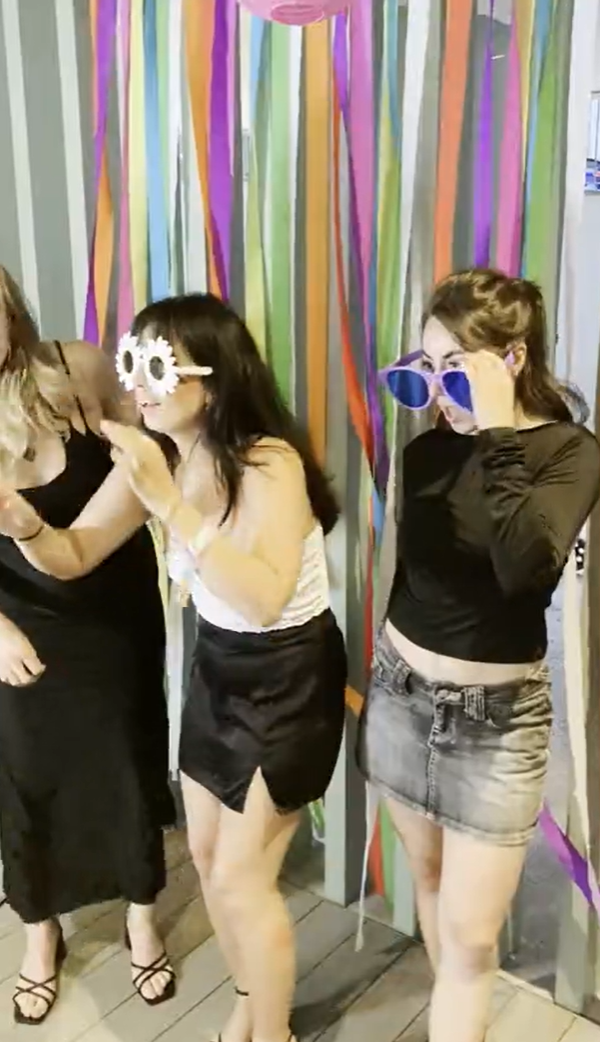 Three people pose playfully in front of colorful streamer decorations. Two wear funky sunglasses, and all are dressed in black and white outfits. They appear to be having fun and enjoying a festive atmosphere.