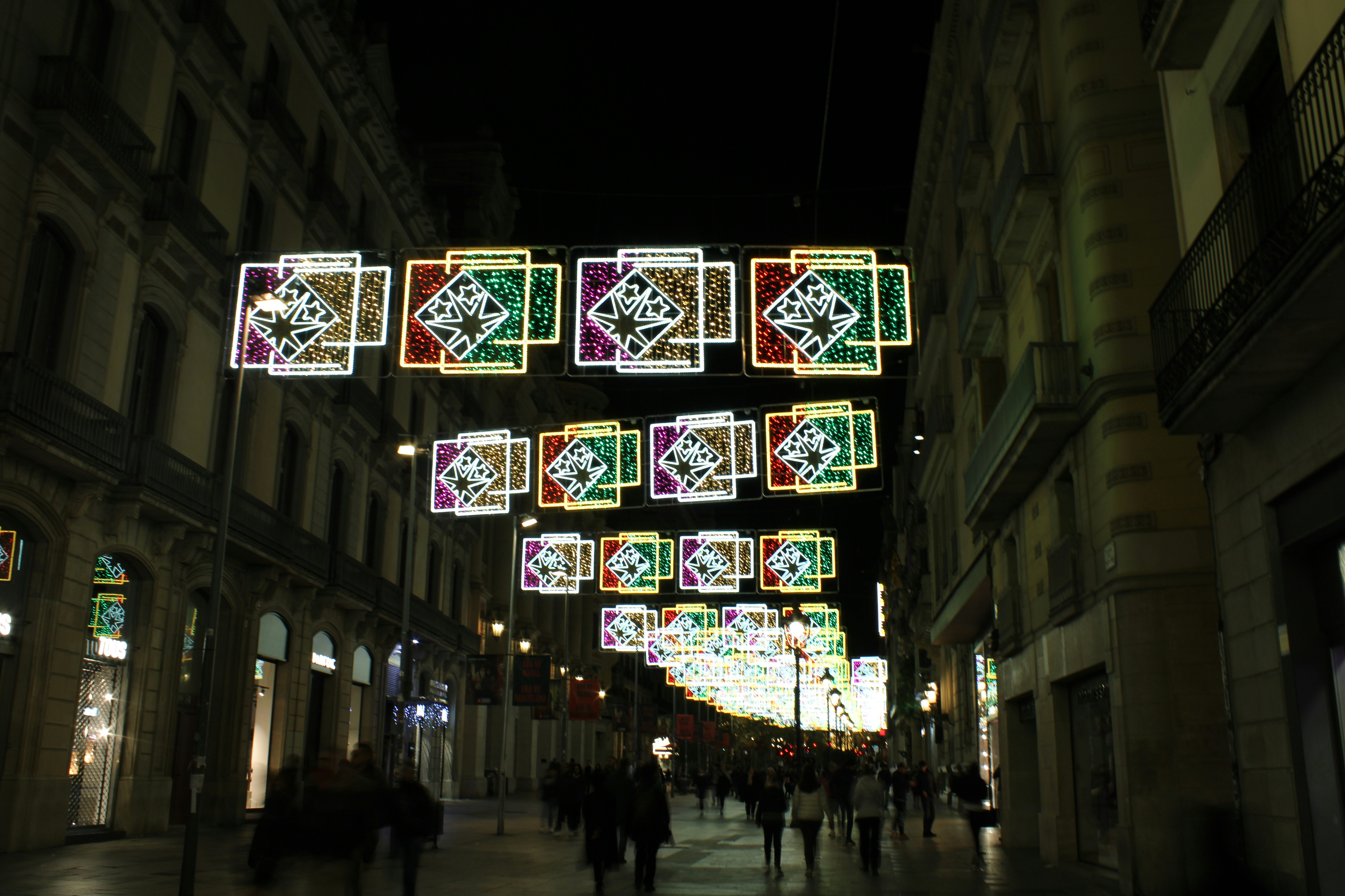 A nighttime street scene in Barcelona with colorful, geometric holiday lights hanging overhead. The luminous squares illuminate the busy street below, where people, including estudiantes making planes navideños, stroll past buildings lining both sides of the street.