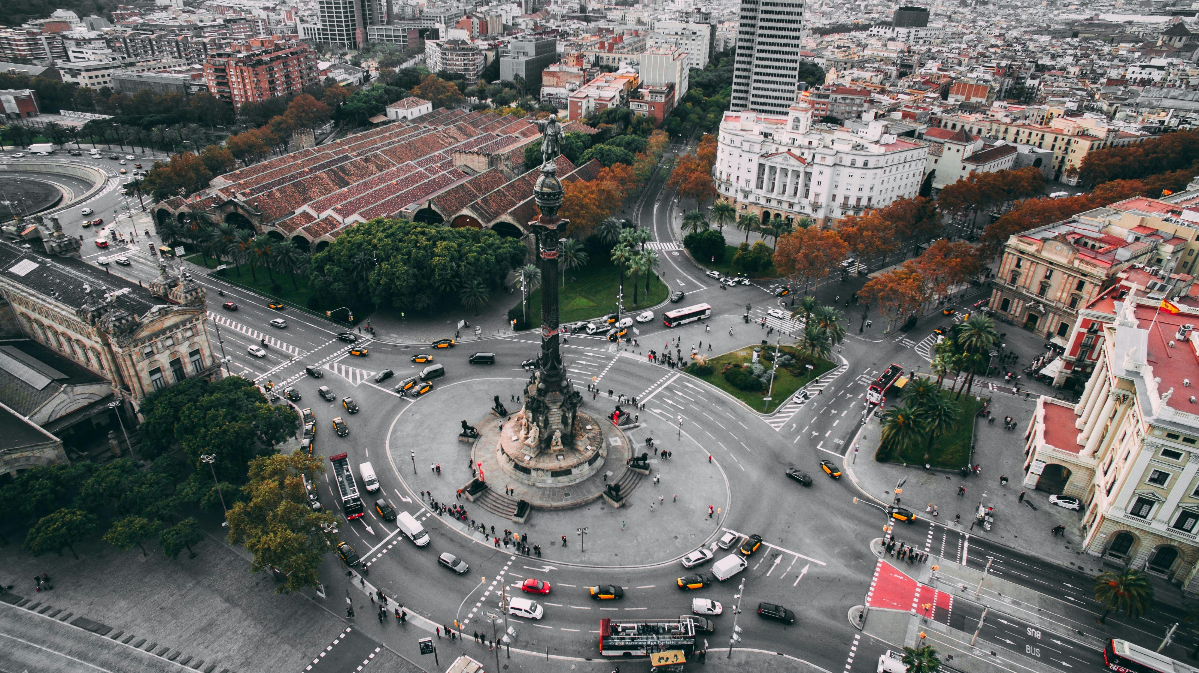 Aerial view of a bustling city roundabout in Barcelona featuring a tall monument in the center, surrounded by traffic and green spaces. Buildings with red roofs and lush trees, some with autumn leaves, enclose the area, creating a vibrant urban scene that captivates both locals and estudiantes.