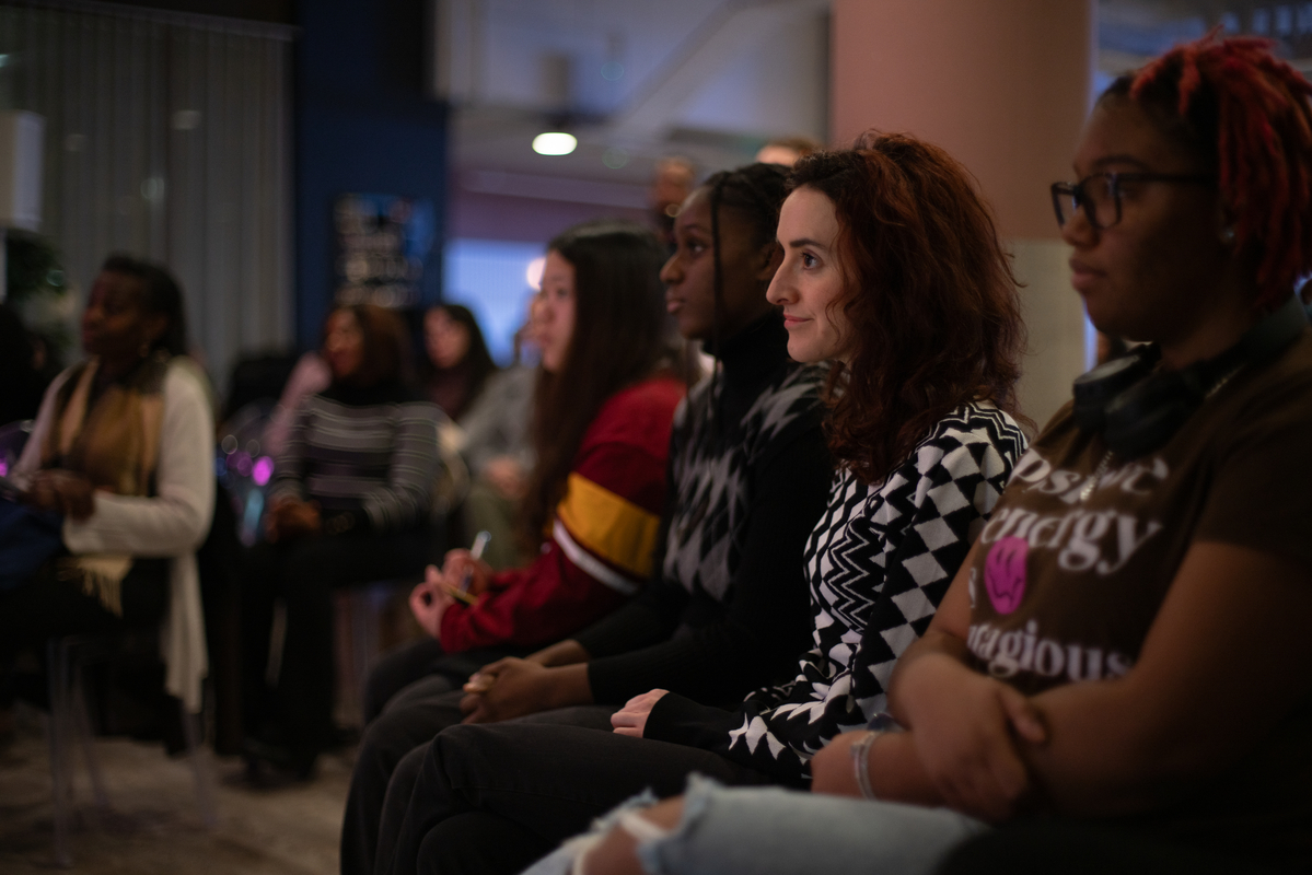 A group of people sit attentively in a dimly lit room. They appear engaged, looking towards something off-camera. The individuals have diverse appearances and are wearing casual clothing. Background elements include other seated people and a large pillar.