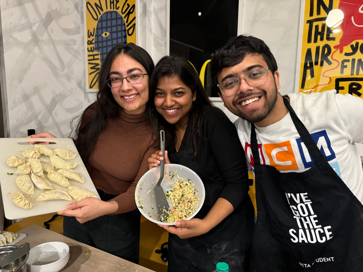 Three people smiling in a kitchen. One person holds a tray of dumplings, another holds a bowl with filling and a spatula, and the third wears an apron with text. Posters hang on the wall behind them.