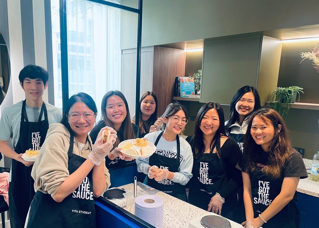 A group of eight people smiling behind a counter, wearing aprons. They are holding pastries and appear to be in a kitchen or cafe setting, with a scale and tissue roll on the counter in front of them.