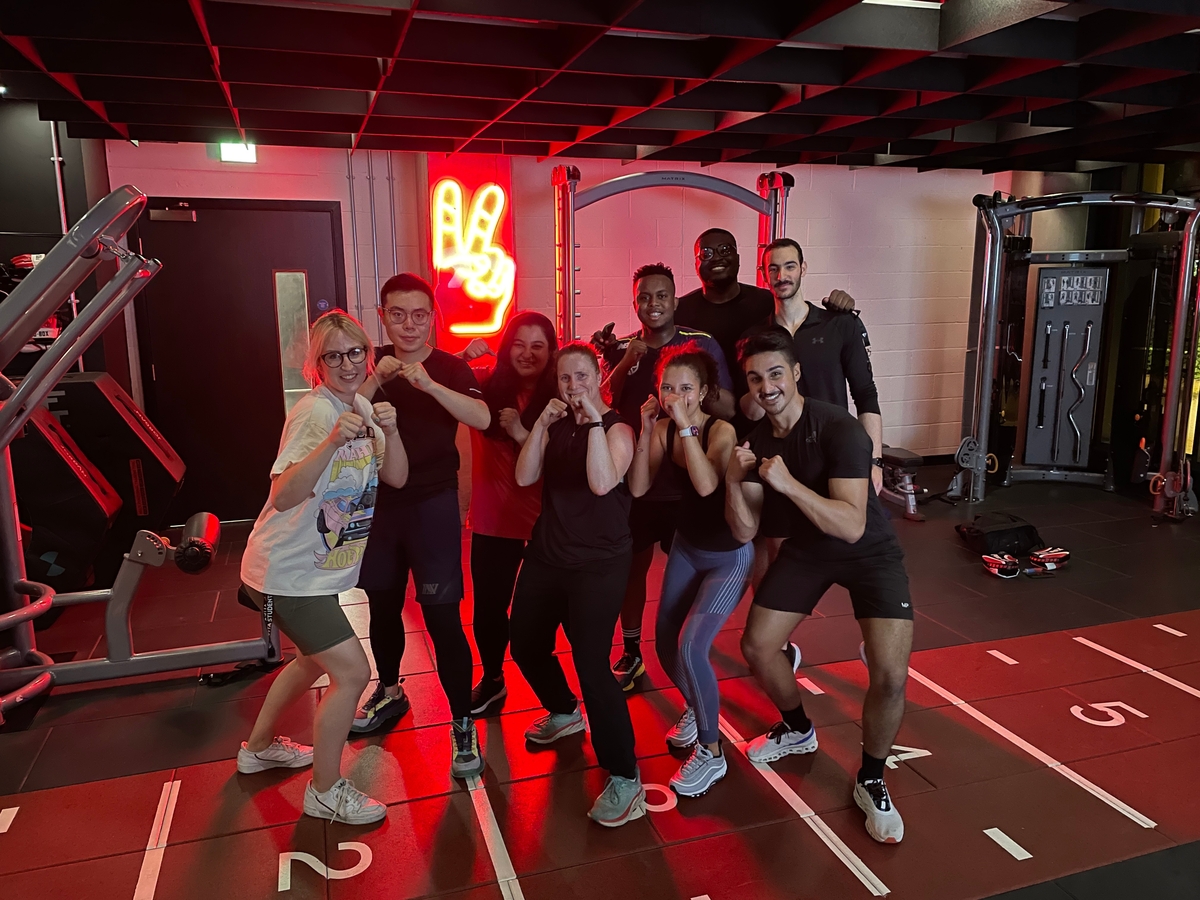 A group of eight people posing in a gym, smiling and holding fists up playfully. They're standing on a marked gym floor with various equipment around. A neon peace sign is lit on the wall behind them.