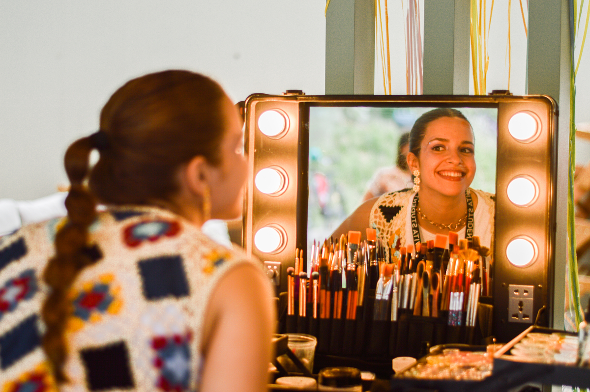 A person with long braided hair and a colorful patterned top smiles in a lit-up makeup mirror. The mirror reflects makeup brushes and cosmetic tools neatly arranged in front of them.