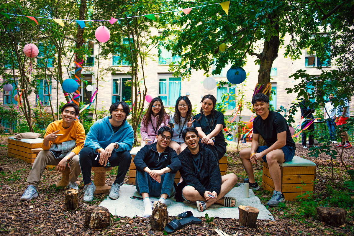 A group of eight people sitting together outdoors on benches and the ground, surrounded by trees and colorful hanging decorations. They are smiling, with some making peace signs. It looks festive, with balloons and lanterns.