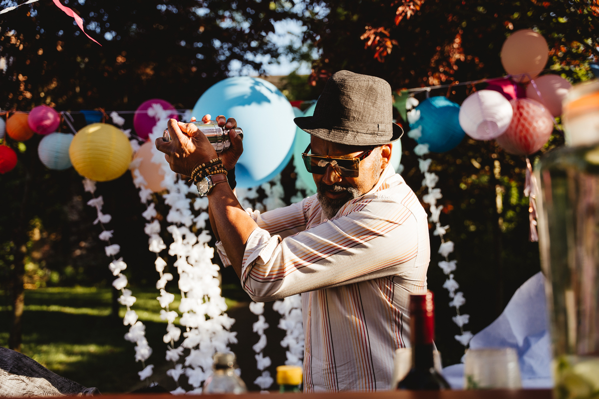 A man wearing a hat and sunglasses shakes a cocktail shaker at an outdoor party. Colorful paper lanterns and white garlands decorate the background, and a bottle of red wine is on the table in front of him. Sunlight filters through the trees.